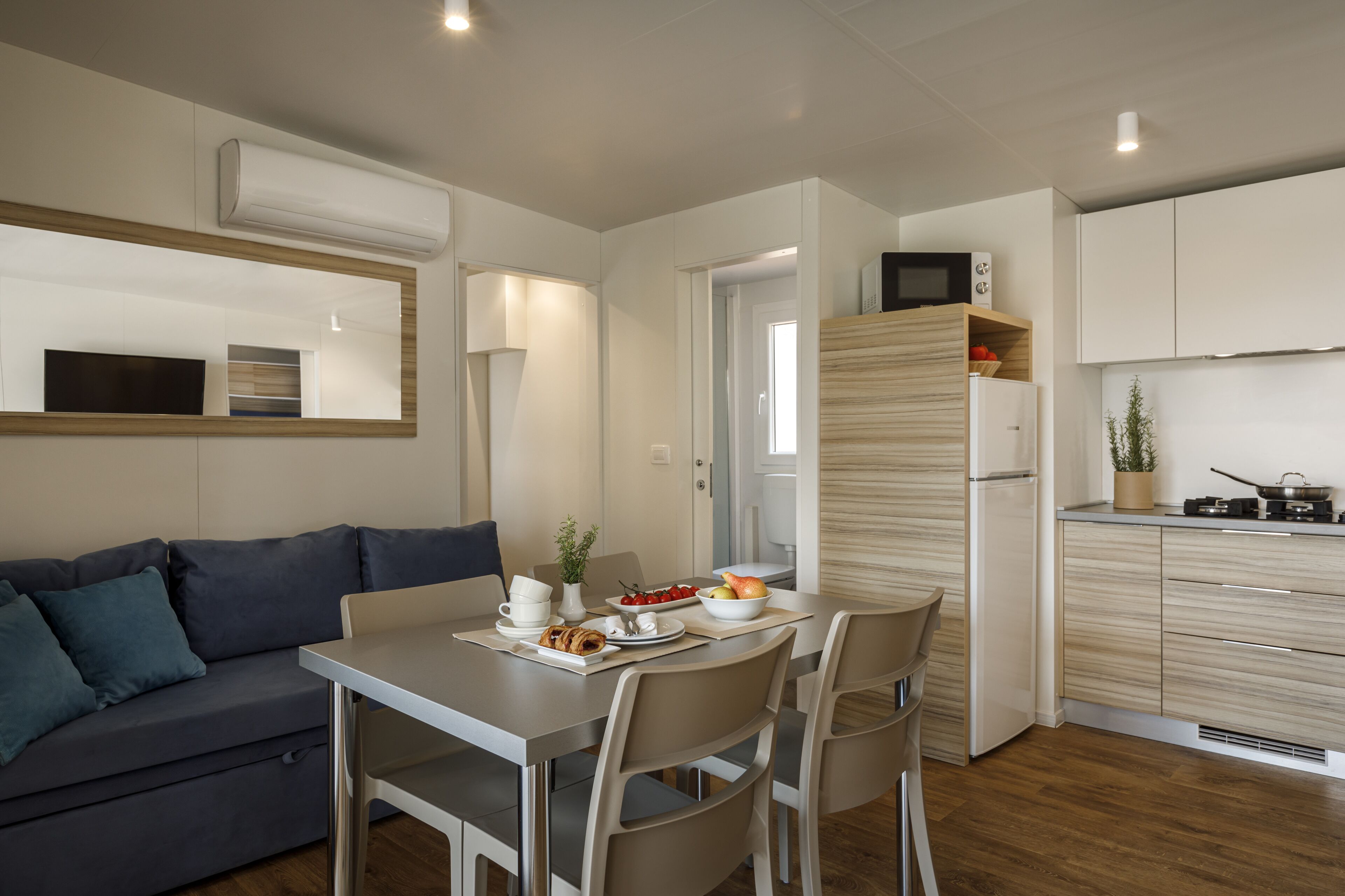 A modern kitchen with white cabinets, a built-in microwave, and a refrigerator. There is a dining table with chairs and a small plant on the counter.