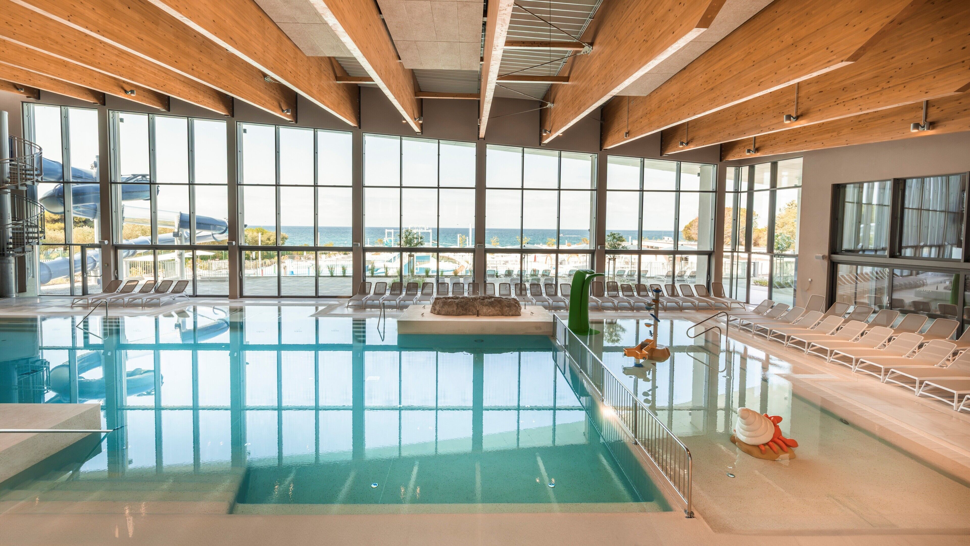Una piscina interior con vistas despejadas al mar a través de amplios ventanales, rodeada de sillones de descanso.
