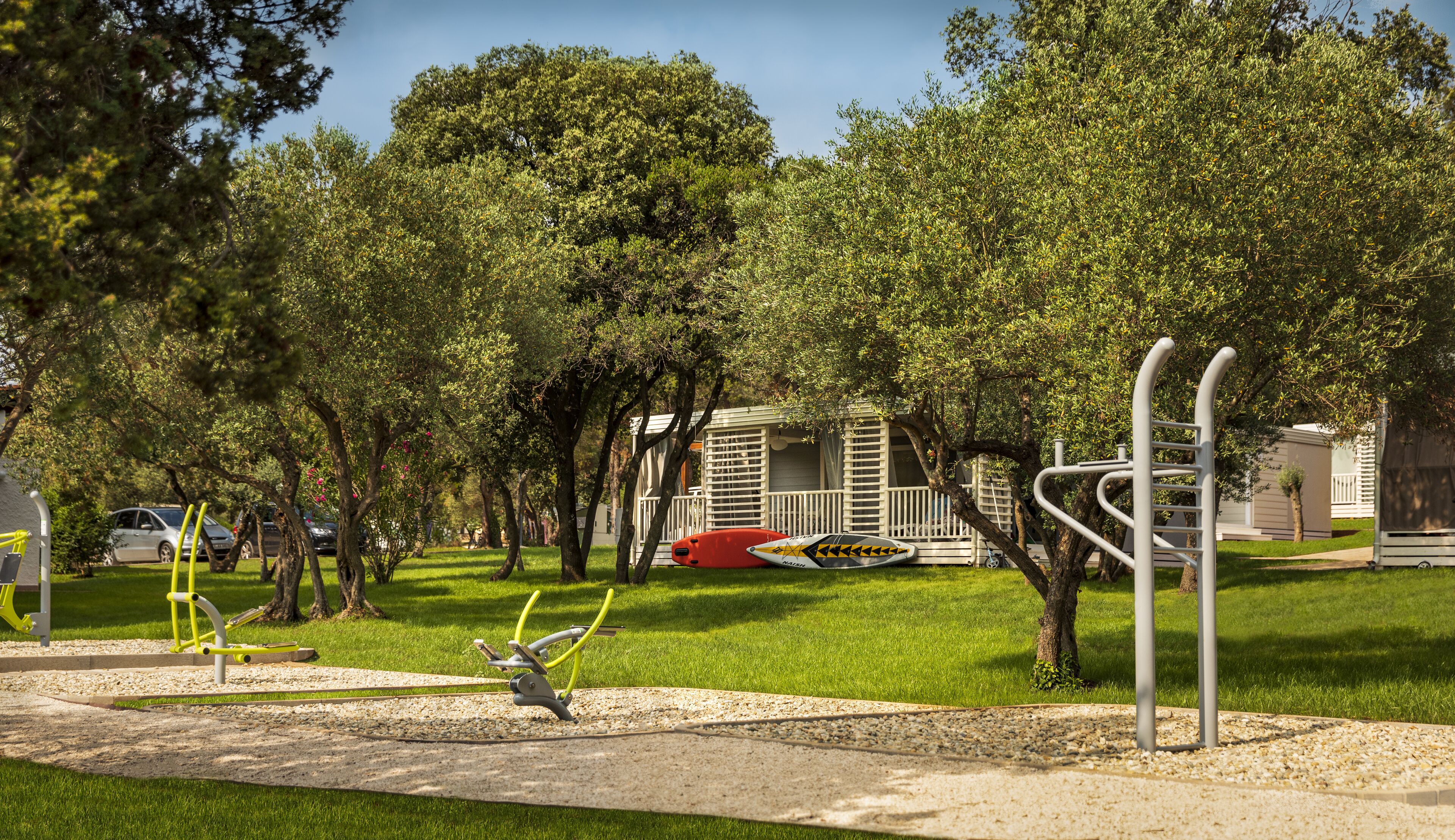 A park with exercise equipment, trees, and mobile homes in the background.