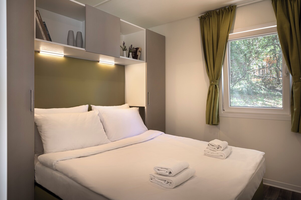 A neatly made bed with white linens and folded towels, a shelf with decorative items, and a window with curtains.