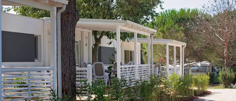 A covered outdoor seating area with white railings and chairs, surrounded by greenery and trees.