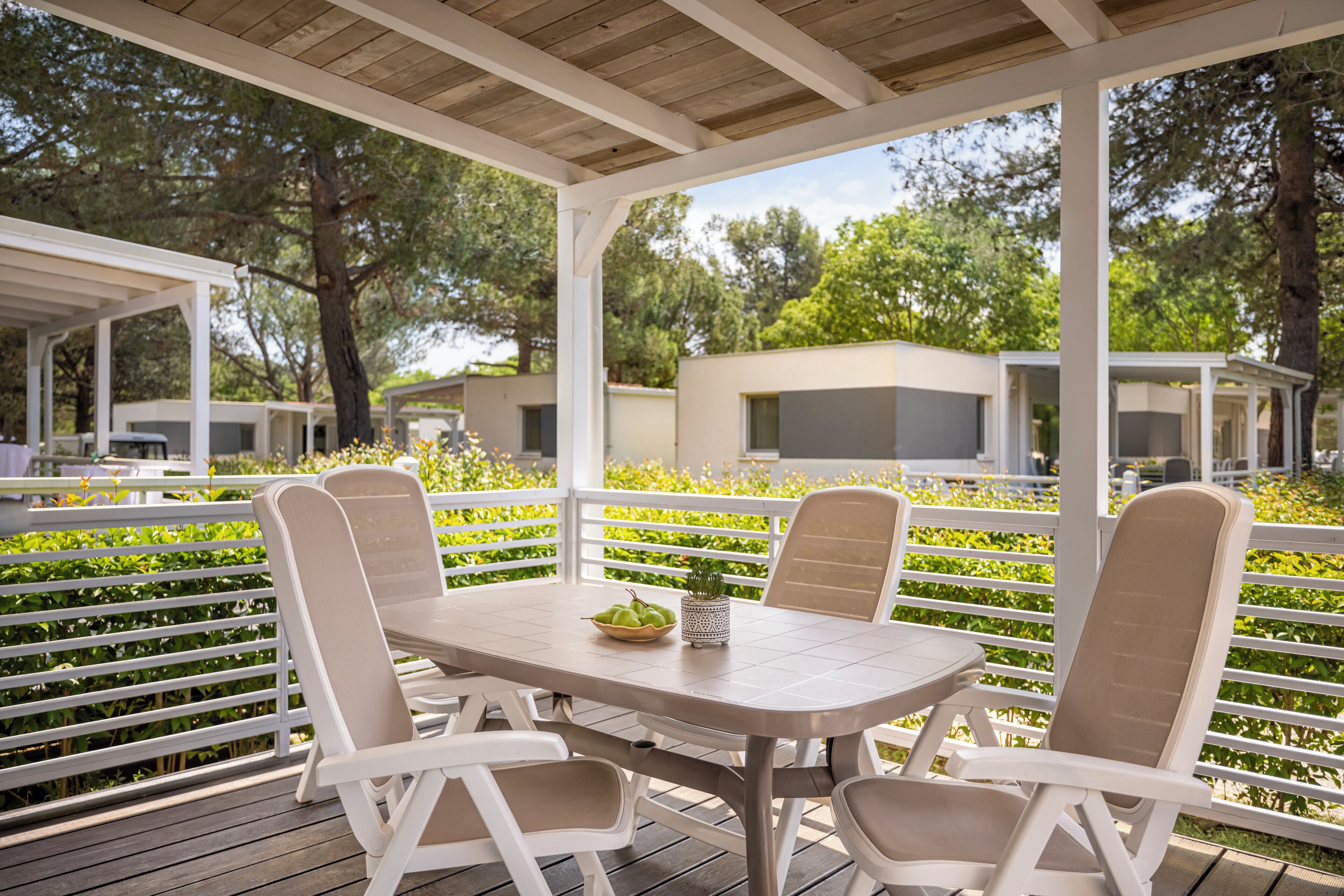 A covered outdoor patio with a table and chairs, surrounded by a white fence and greenery.