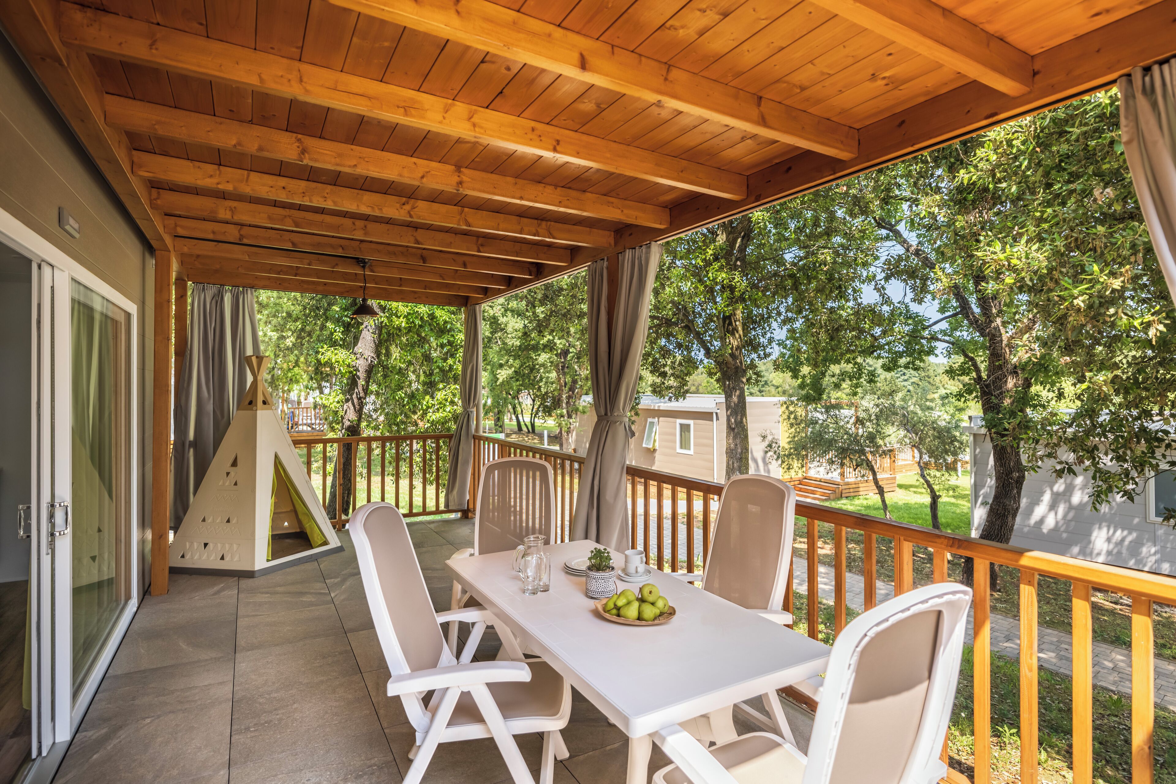 Een overdekt terras met een witte tafel en stoelen, een speelstructuur voor kinderen en uitzicht op de omliggende bomen en huizen.