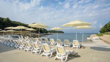 A beachside lounge area with rows of white lounge chairs and beige umbrellas, overlooking the sea.