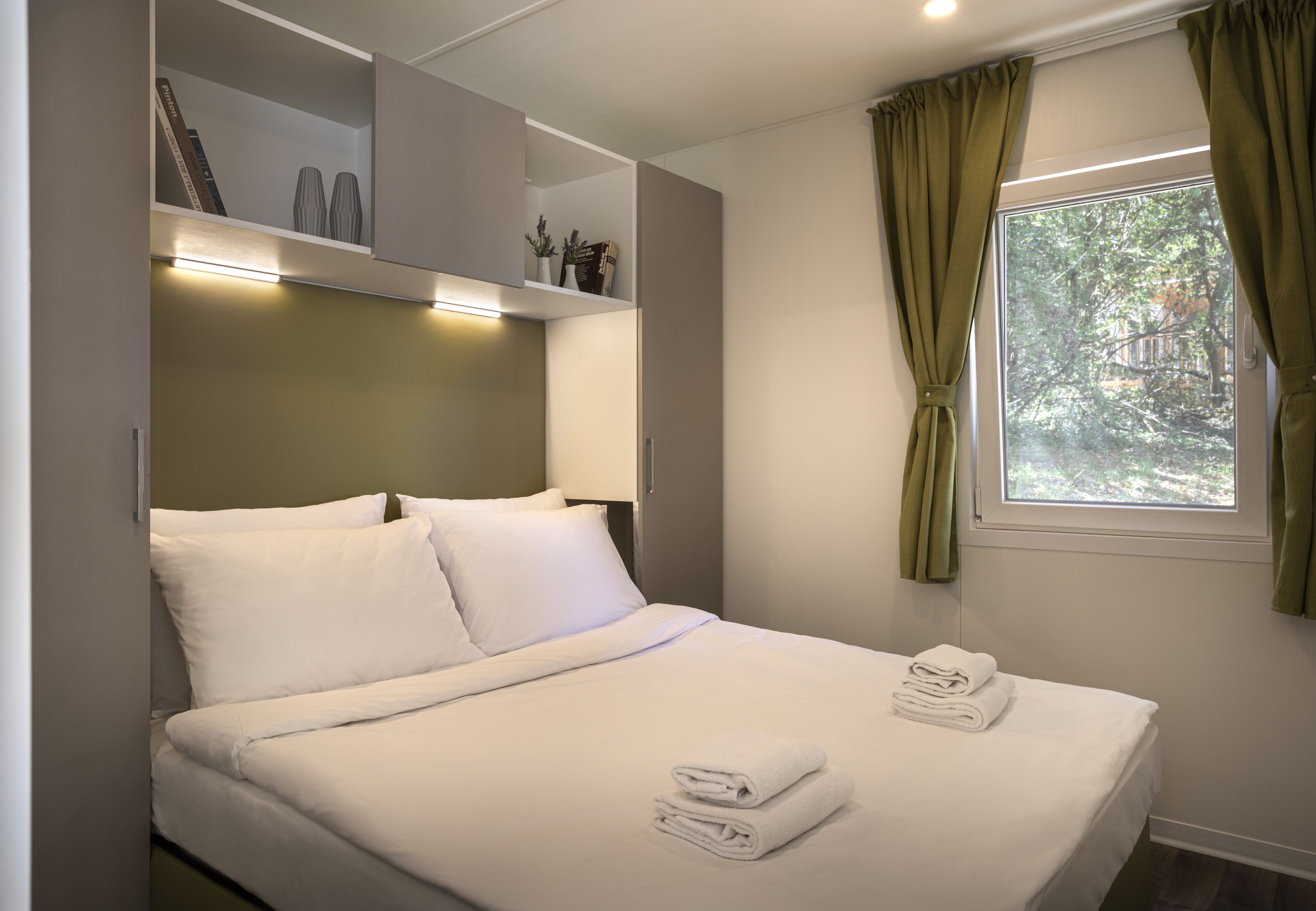 Un lit bien fait, avec du linge de lit blanc et des serviettes pliées, une étagère ornée d’objets décoratifs et une fenêtre avec des rideaux.
