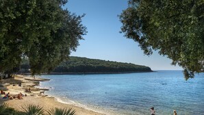 A beach with people relaxing, clear blue water, and a forested hill in the background.