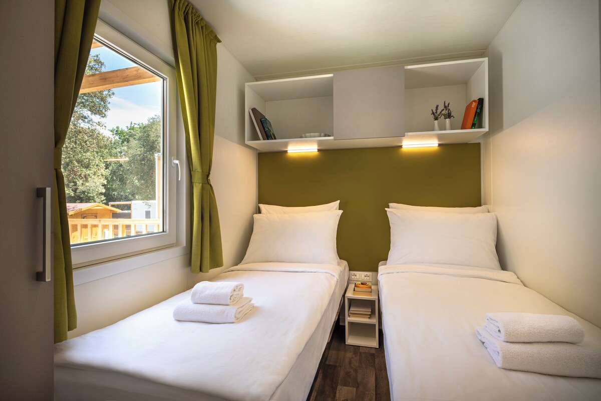 A hotel room with two beds, a window with curtains, a shelf with books and a vase, and a nightstand with a lamp.