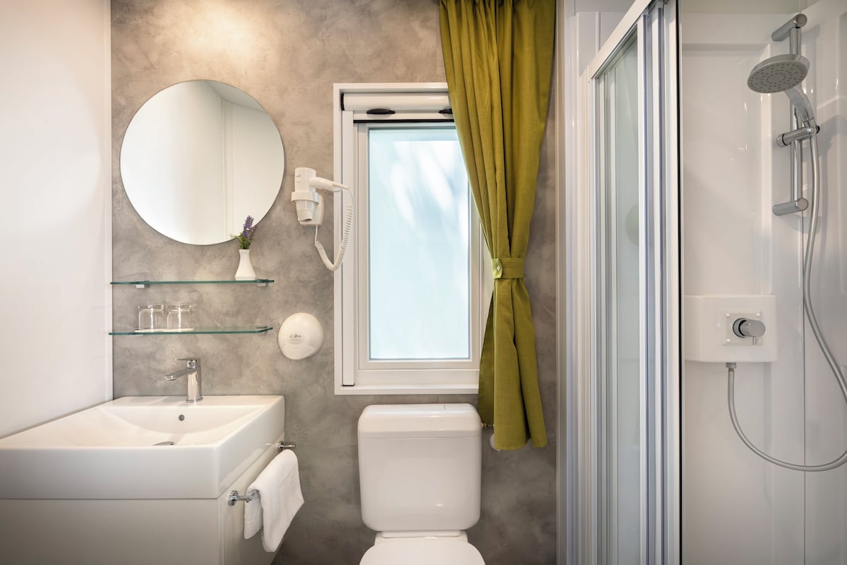 A bathroom with a white sink, a toilet, a glass shelf with toiletries, a round mirror, and a shower with a glass door.