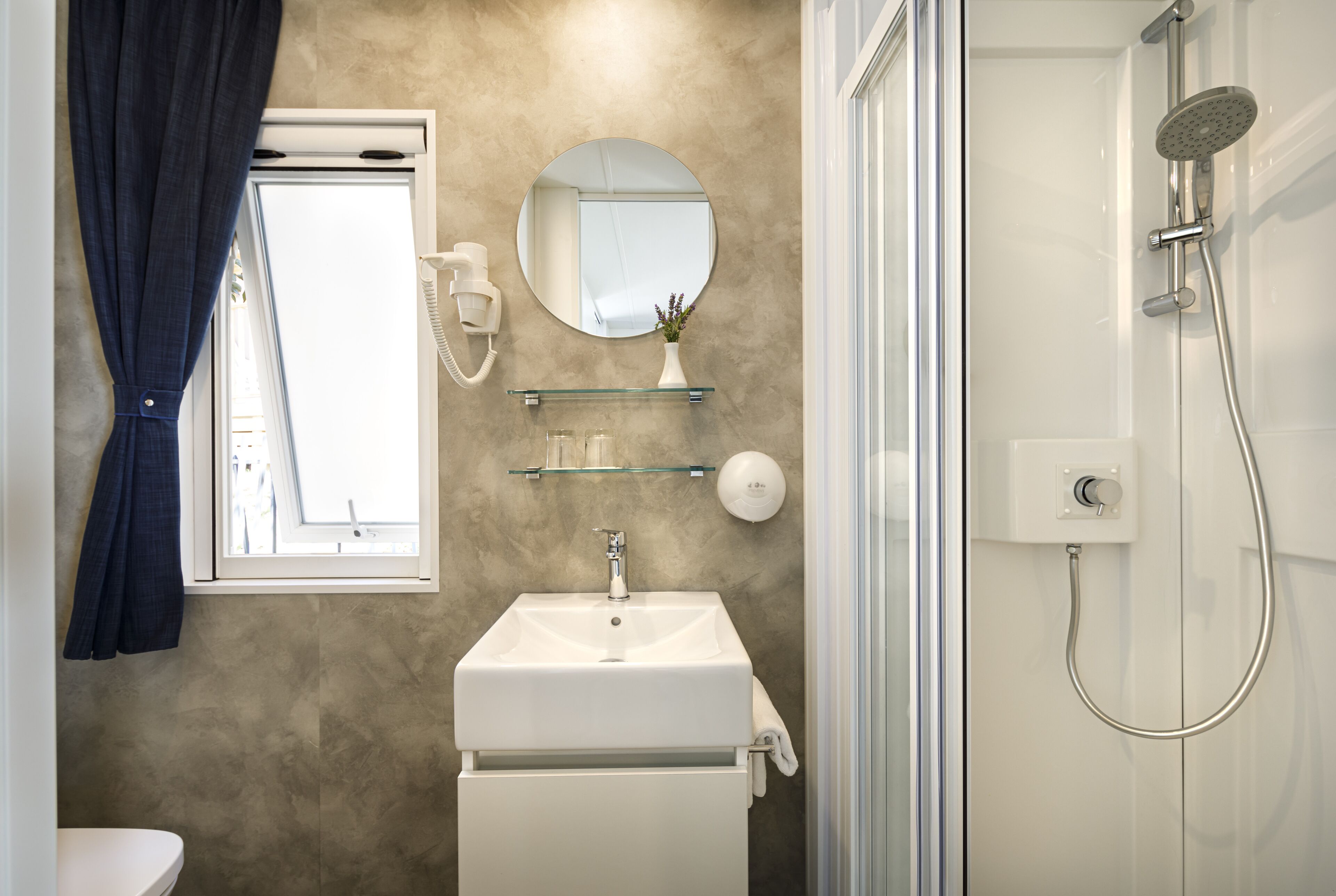 Une salle de bain avec un lavabo blanc, un miroir, une étagère en verre, une douche et une fenêtre avec des rideaux.