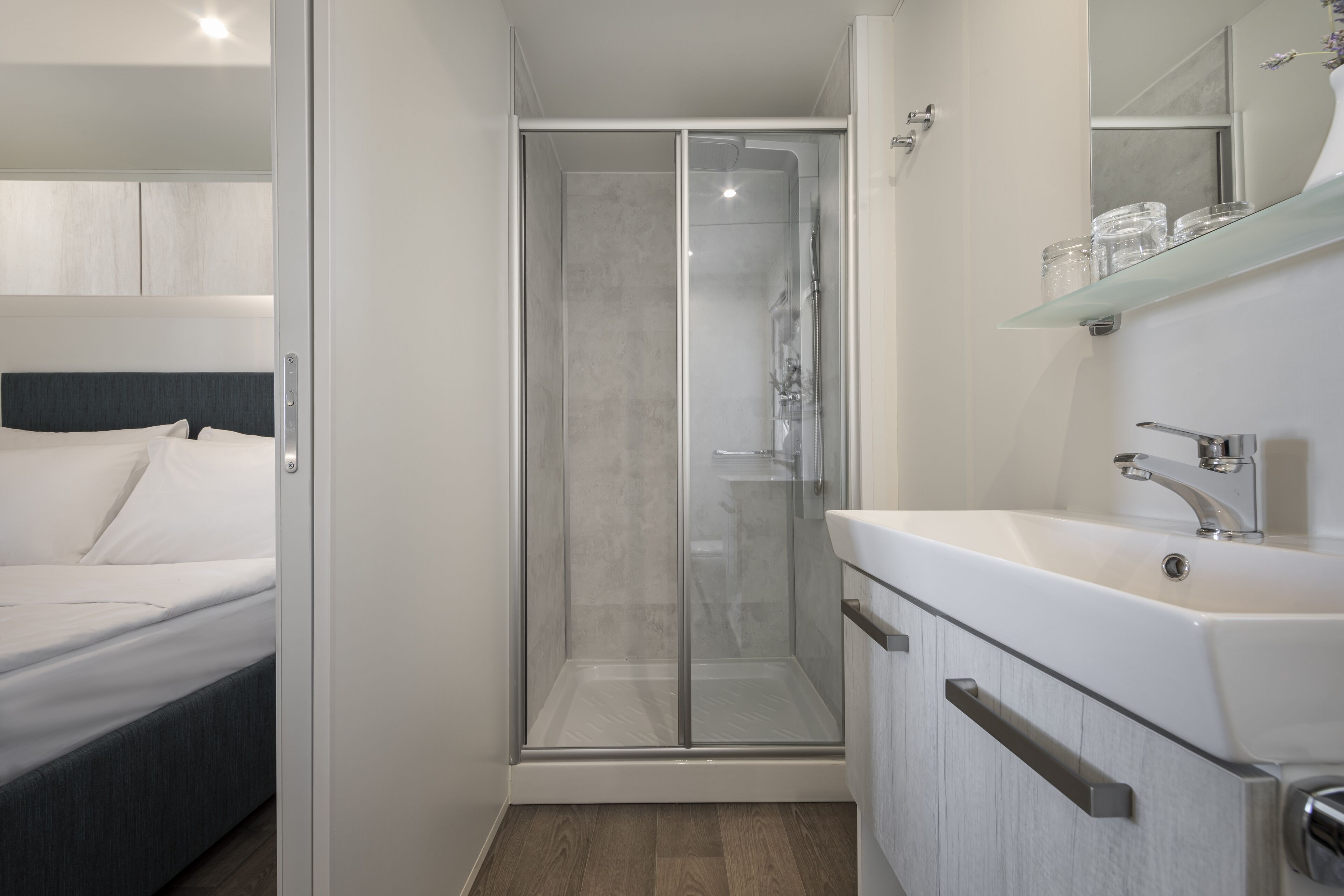 A modern bathroom with a glass shower enclosure, a white sink, and a bedside table with a bed.