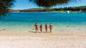 A beach with clear blue water, a sandy shore, and a few people walking.