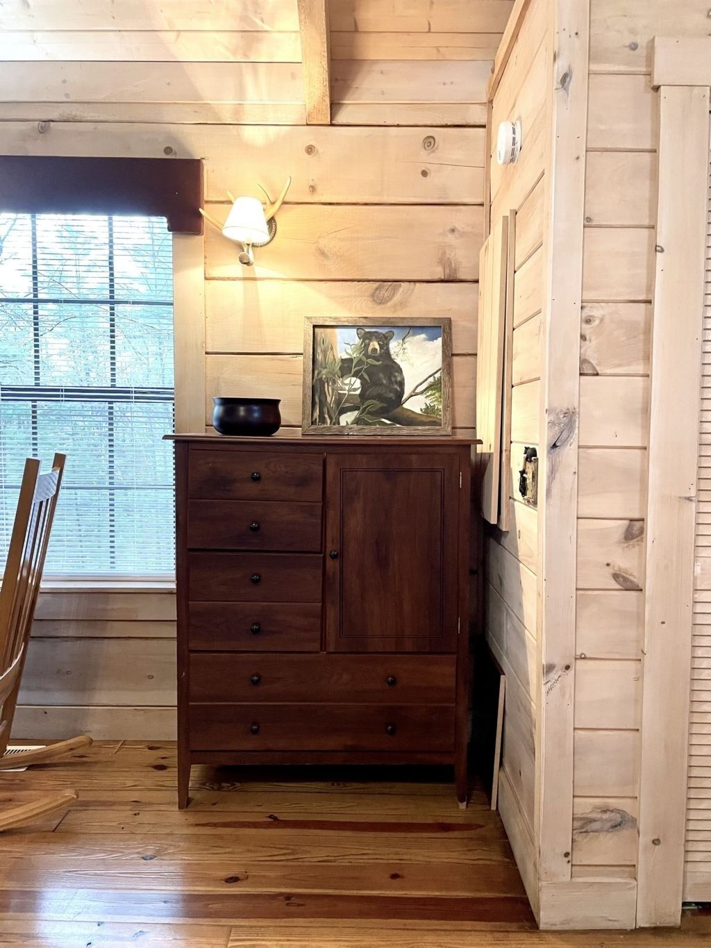 A wooden cabin interior with a wooden cabinet, a framed picture of a bear, a lamp, and a window with blinds.
