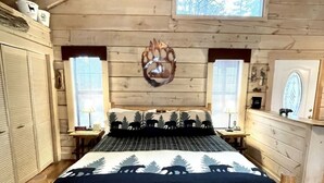 Une chambre de style cabane en bois, avec un lit orné d’un motif représentant un ours et un arbre, une tête de lit en bois et une petite fenêtre située au-dessus du lit.