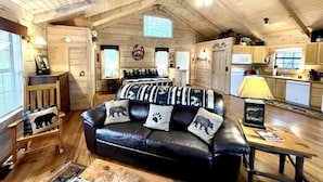 L’intérieur d’un chalet accueillant, avec un canapé en cuir noir, des murs en bois, un ventilateur de plafond et un coin cuisine.