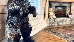 Une sculpture d’ours en bois, avec l’inscription « BIENVENUE » sur une base en bois, dans une pièce aménagée dans le style d’un chalet, avec une cheminée et un téléviseur.