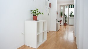 A bright hallway with white walls, a wooden floor, and a white shelving unit with a potted plant. There are doors and windows along the hallway, and a view of a balcony with plants. - Marimari Apartment, 4th floor with elevator (Split)