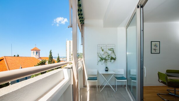 A balcony with a white railing, a small table with two chairs, a potted plant, and a sliding glass door leading to a room with a green chair and a framed picture on the wall. - Marimari Apartment, 4th floor with elevator (Split)