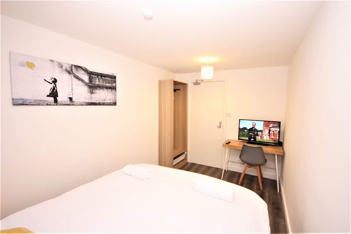 A hotel room with a bed, a desk with a computer, a painting on the wall, and a door leading to another room.
