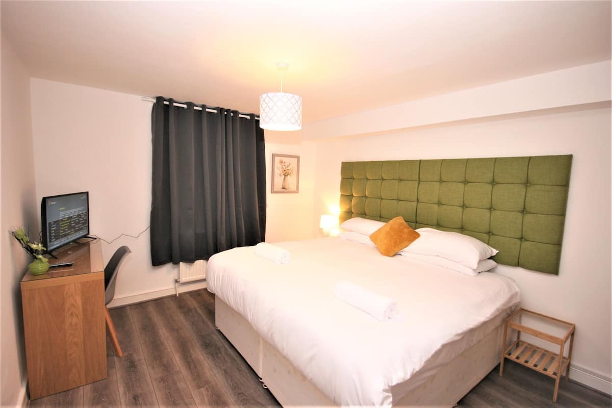 A hotel room with a bed, a desk with a computer, a chair, a small table, and a window with curtains.