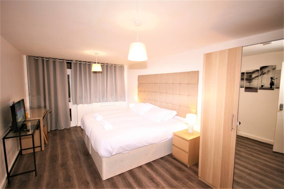 A hotel room with a large bed, a desk with a computer, a bedside table with a lamp, and a wardrobe.