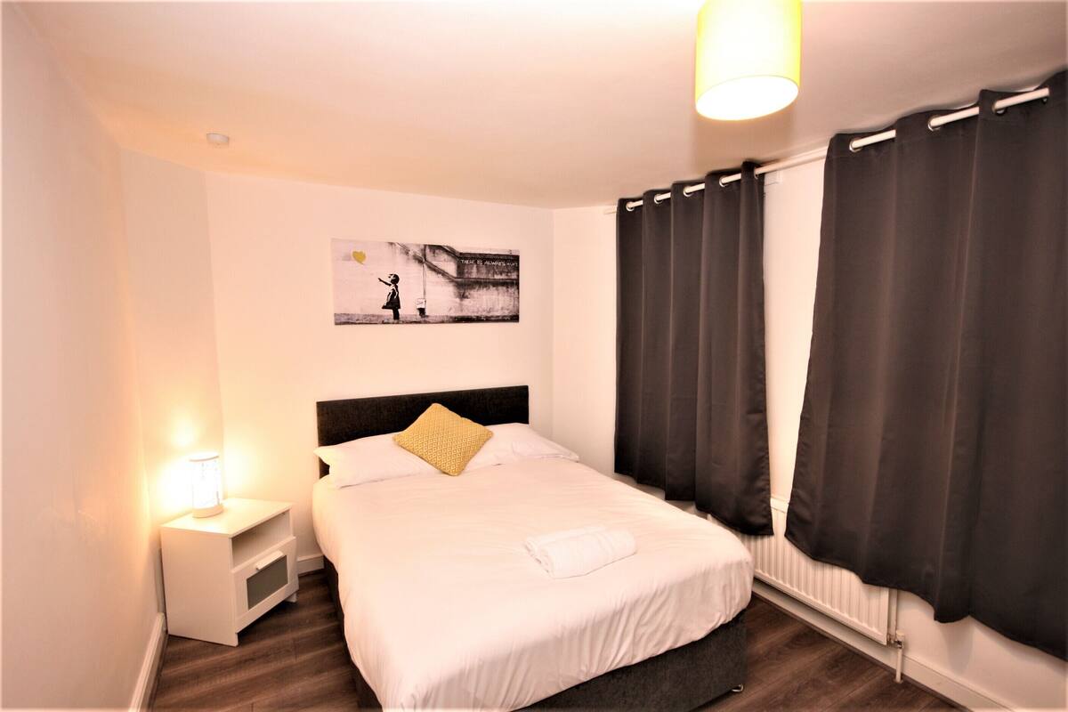 A neatly arranged hotel room with a bed, a nightstand, a lamp, a painting on the wall, and dark curtains.
