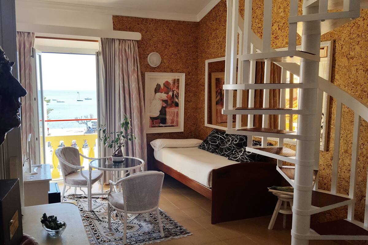 Une pièce comprenant un lit, un canapé, un escalier, un coin repas avec une table et des chaises, et une vue sur la mer par une grande fenêtre.