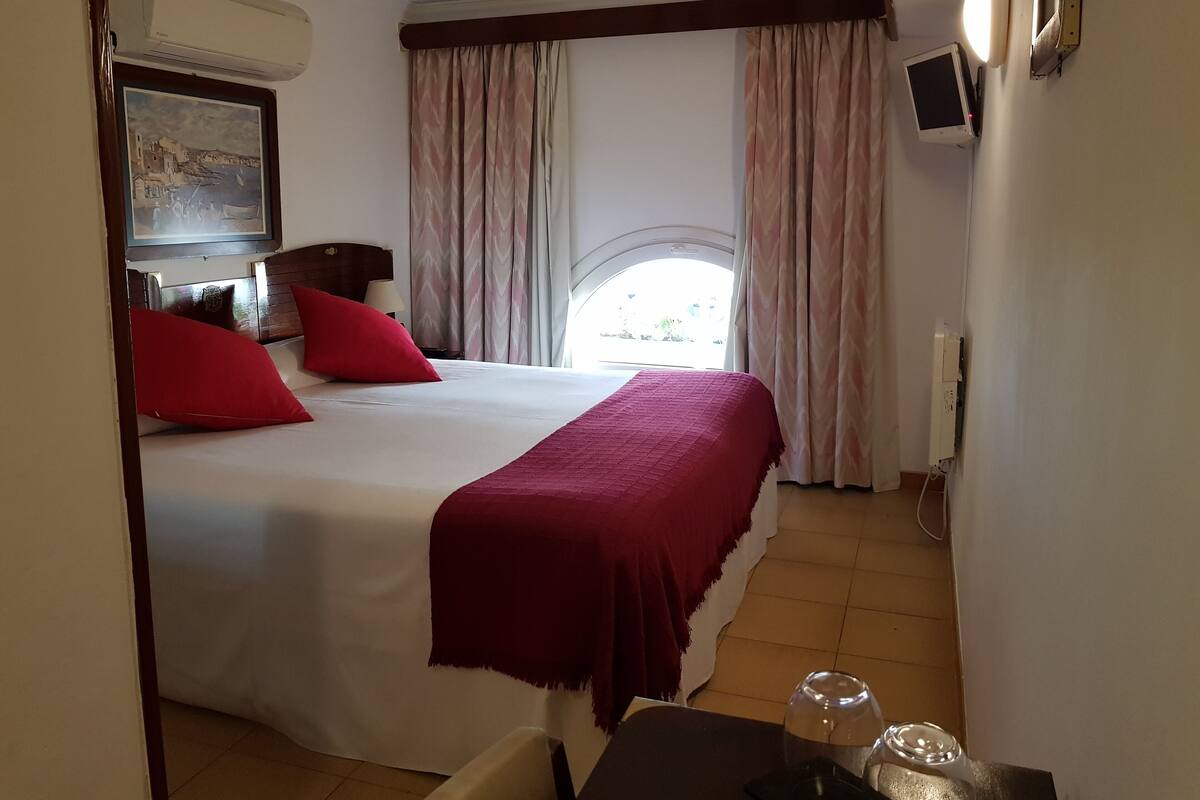 Une chambre d’hôtel comprenant un lit, un bureau, une chaise, une fenêtre avec des rideaux, un téléviseur fixé au mur et un appareil de climatisation.