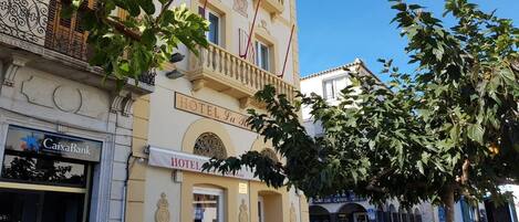 La façade d’un hôtel, avec une enseigne indiquant « HÔTEL La Riviere », une banque nommée « CaixaBank » sur la gauche, et une rue bordée de piétons et d’arbres.