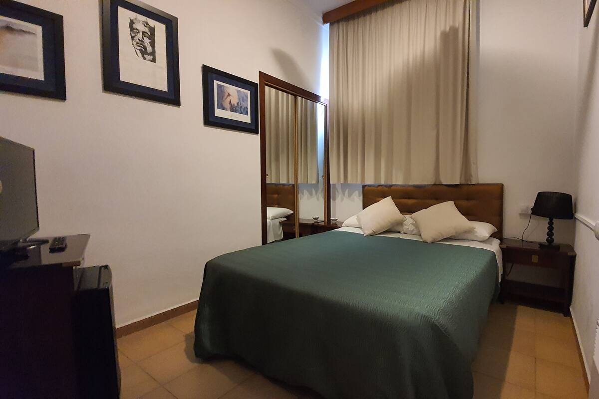 Une chambre d’hôtel avec un lit, une table de chevet, une lampe, un téléviseur et des tableaux encadrés au mur.