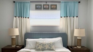A bedroom with a bed, two nightstands, a window with curtains, and a wall with framed pictures.