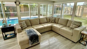 A large L-shaped beige sectional sofa with a throw blanket, a small black side table with magazines, and a large window overlooking a backyard with a pool.
