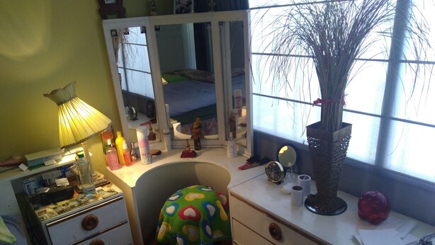 Een kleine slaapkamer met een bed, een bureau met lamp, een spiegel en een raam met uitzicht op bomen.