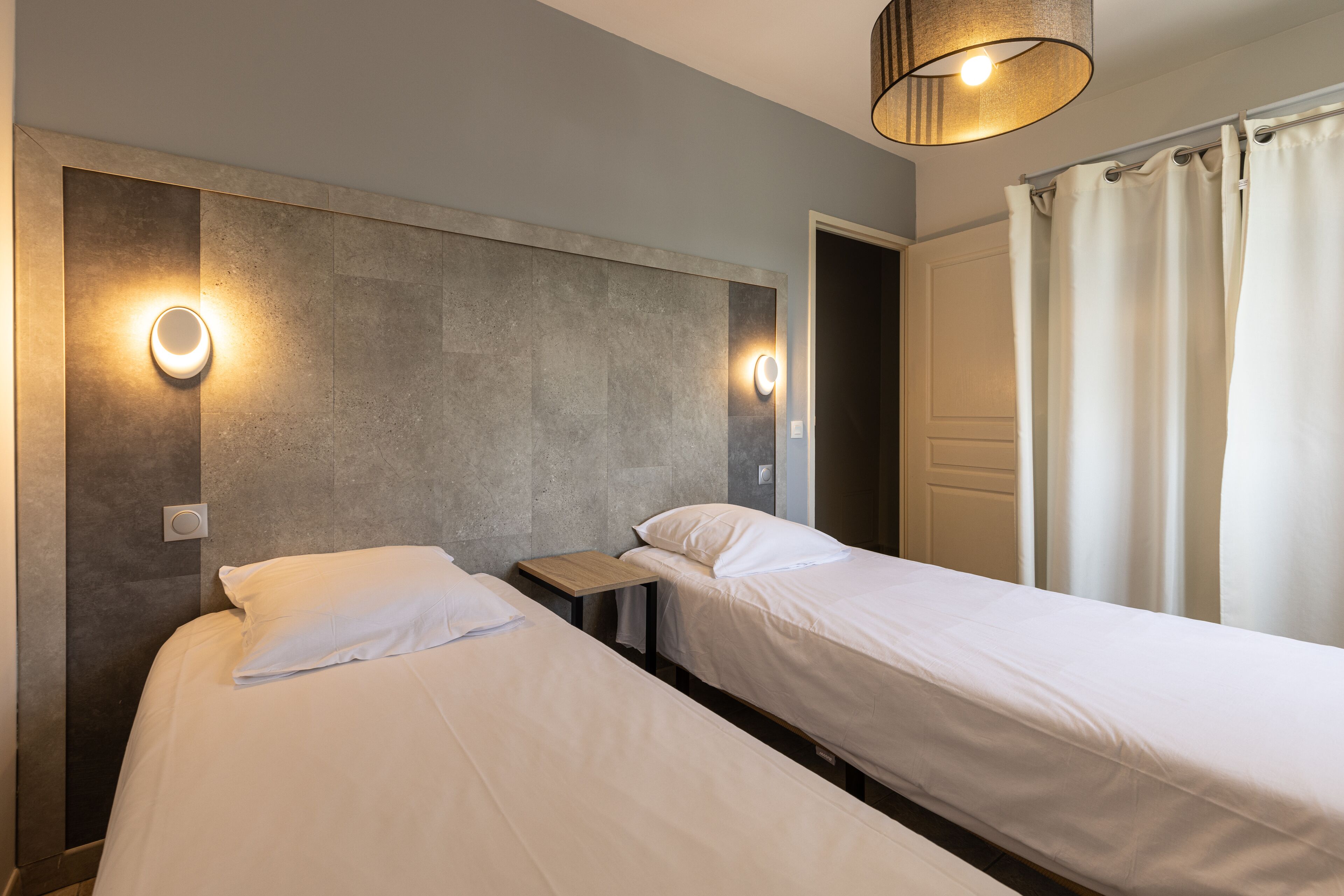 A hotel room with two single beds, a small wooden nightstand, and two wall-mounted light fixtures.