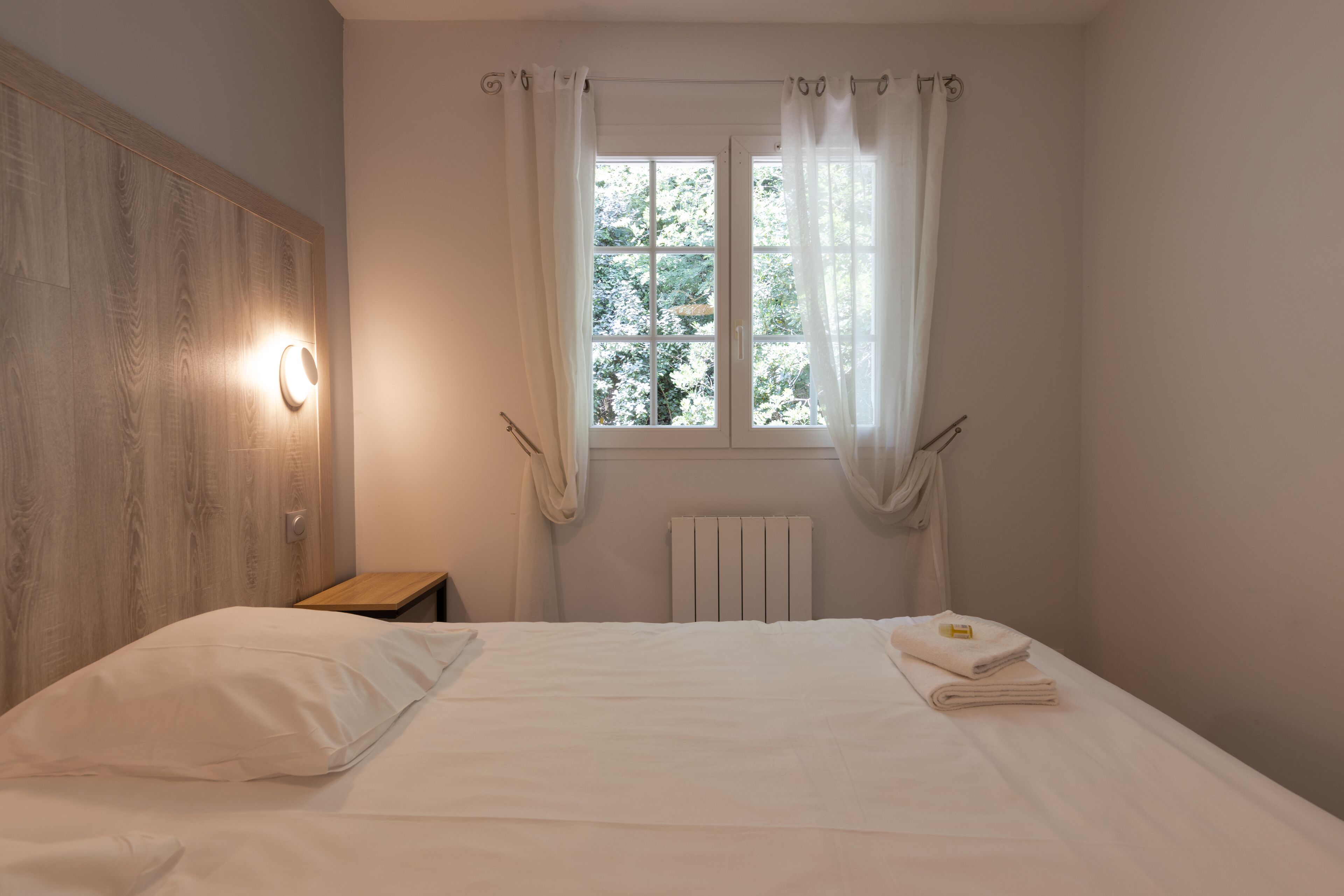 A bedroom with a wooden headboard, a bedside table, a window with sheer curtains, and a radiator.
