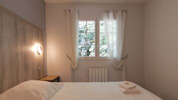 A bedroom with a wooden headboard, a bedside table, a window with sheer curtains, and a radiator.
