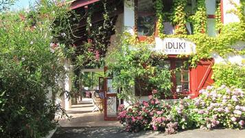 A building with a sign reading "iDUKI OSTAUA" surrounded by lush greenery and pink flowers.