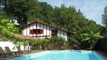 Una piscina con sillones de descanso y una casa con contraventanas rojas.