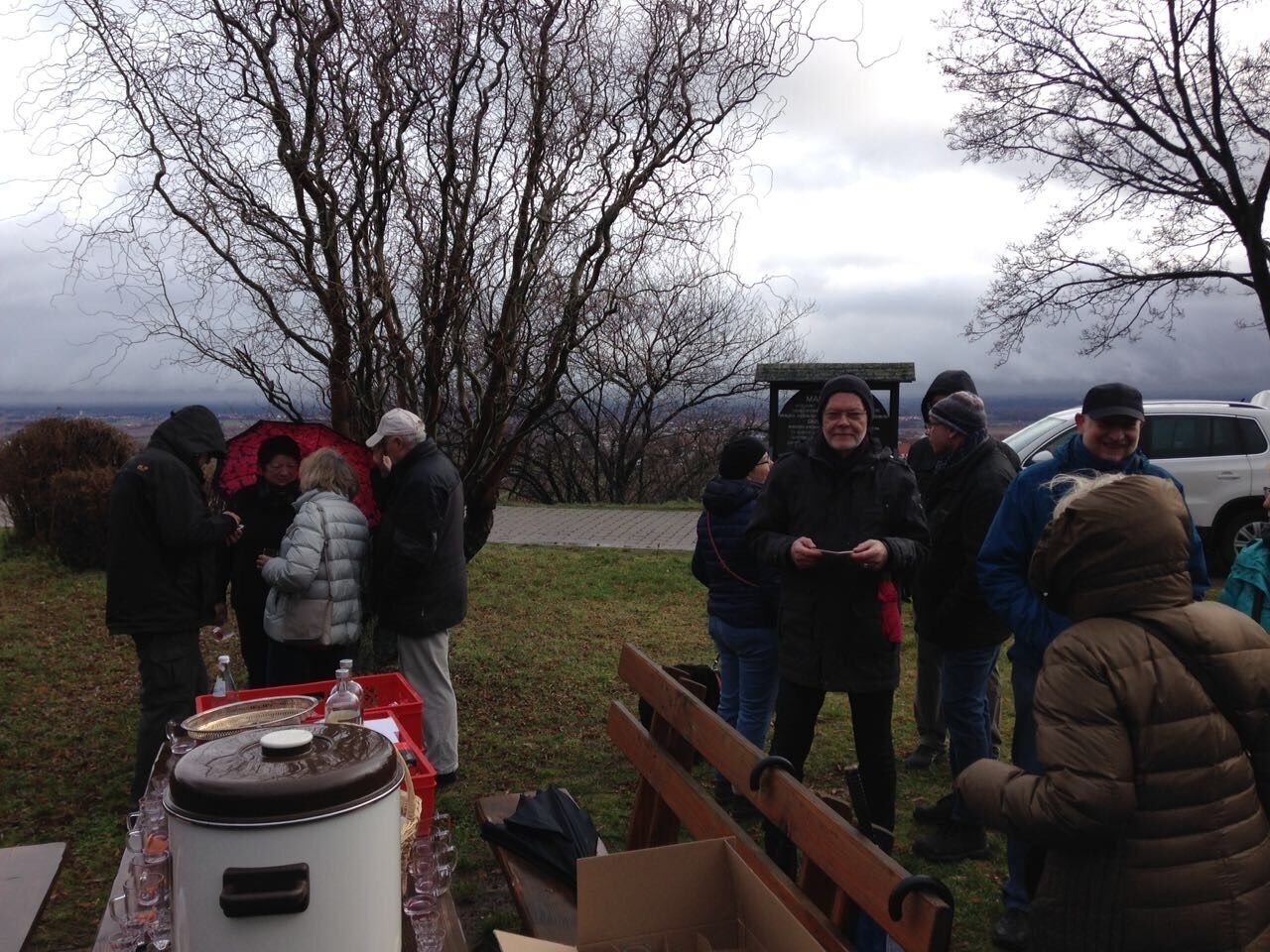 En gruppe mennesker samlet udendørs, måske ved et picnic eller arrangement, med et bord dækket med en stor beholder, en bænk og nogle personlige ejendele.
