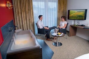 A hotel room with a large bathtub, a desk with a computer, a television displaying a vineyard, and a dining table with fruits and glasses. - Weingut Schreieck VINOTEL & GUTSHAUS (Sankt Martin)