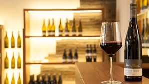 A wine bottle and glass on a wooden counter with a backdrop of wine bottles.