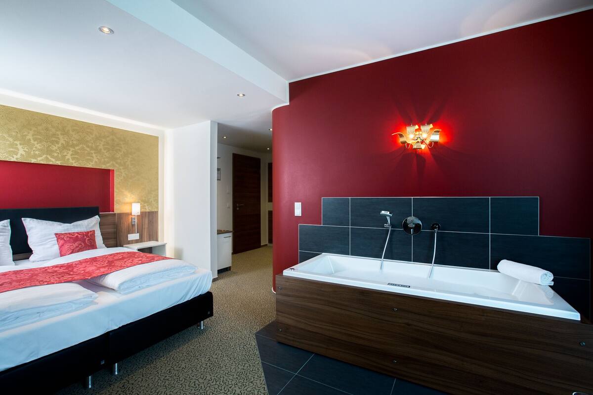 Een moderne hotelkamer met een groot bed, een houten hoofdbord, een rode accentmuur, een wandlamp en een vrijstaand bad.