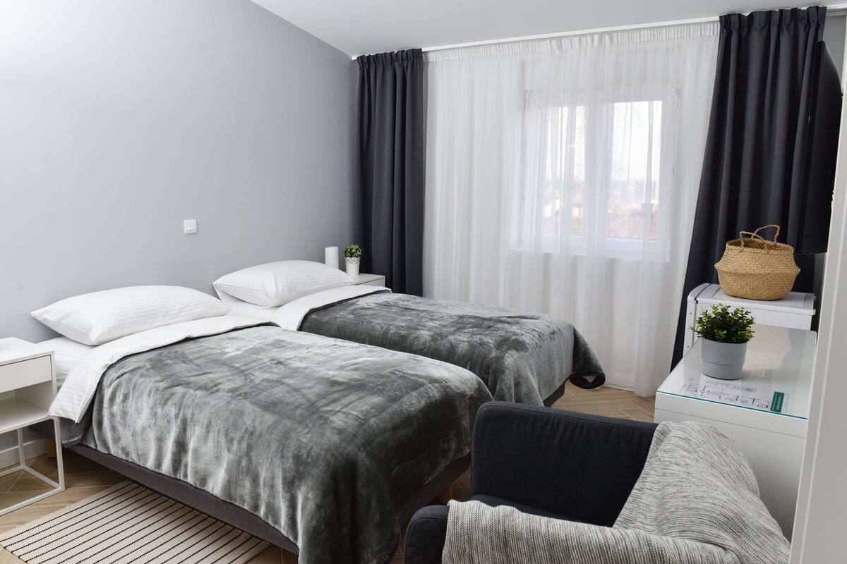 Ein modernes Schlafzimmer mit zwei Betten, einem Nachttisch, einem Sessel und einem Fenster mit Vorhängen.