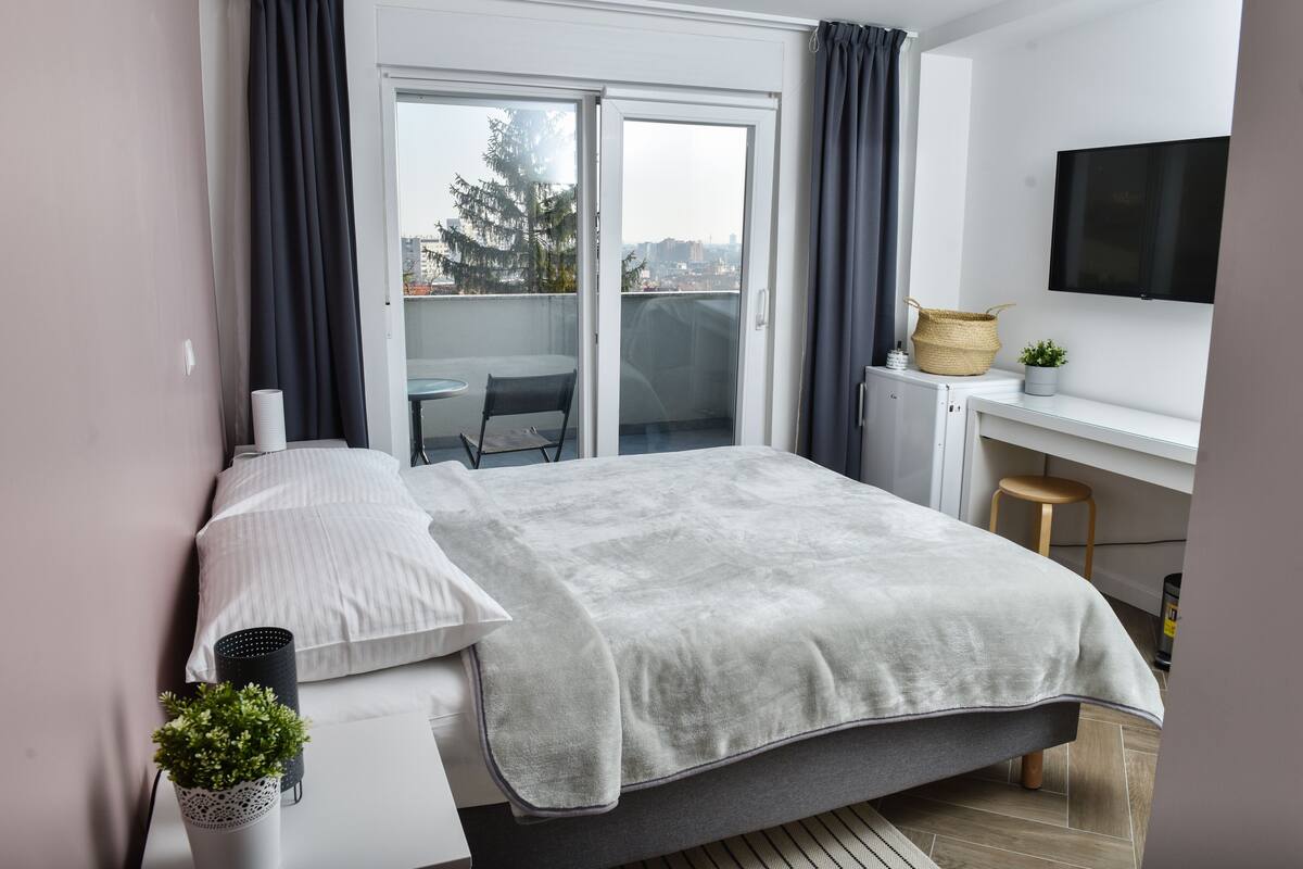 Ein modernes Schlafzimmer mit Bett, Schreibtisch und Blick auf die Stadt durch eine Schiebetür.