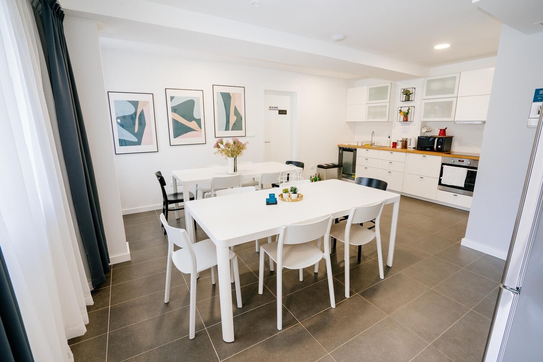Una cocina moderna con armarios blancos, una zona de comedor con sillas blancas y una pequeña mesa. Hay cuadros enmarcados en la pared, un microondas y una cafetera.