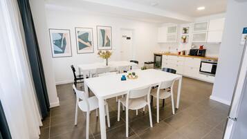 Una cocina moderna con armarios blancos, una zona de comedor con sillas blancas y una pequeña mesa. Hay cuadros enmarcados en la pared, un microondas y una cafetera.