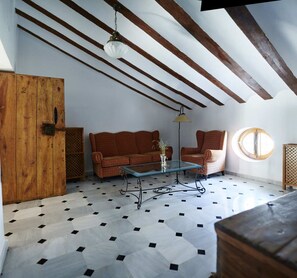 Een kamer met schuine plafonds, houten balken, een patroon vloer, een bank, een stoel, een tafel en een houten deur.