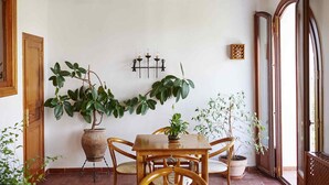 Una habitación con una mesa y sillas de madera, plantas en macetas, una lámpara fijada a la pared y un ventanal grande.