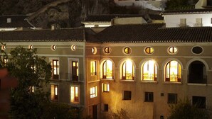 Un edificio con varios balcones y ventanas arqueadas, rodeado de follaje y una ladera rocosa.