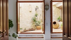 Un pasillo con puertas de madera, suelo de baldosas y vista a un patio con plantas y una maceta grande.