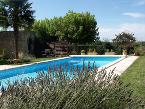 A rectangular swimming pool with a stone wall on one side and a wooden deck on the other. There are chairs and a table under a pergola, surrounded by various plants and trees. - Chambre d'hôtes Les Lavandes available for 3 consecutive nights only (Beaumont-du-Périgord)